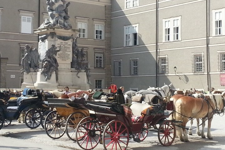 Horse-drawn carriages and statue in a city square with historic buildings in the background.