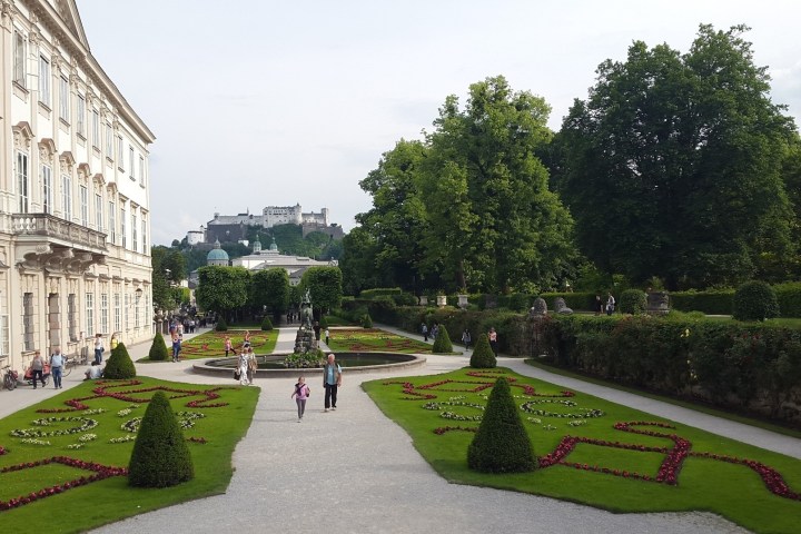 Ornate garden with patterned flower beds, a pathway, and a historic building on the left.