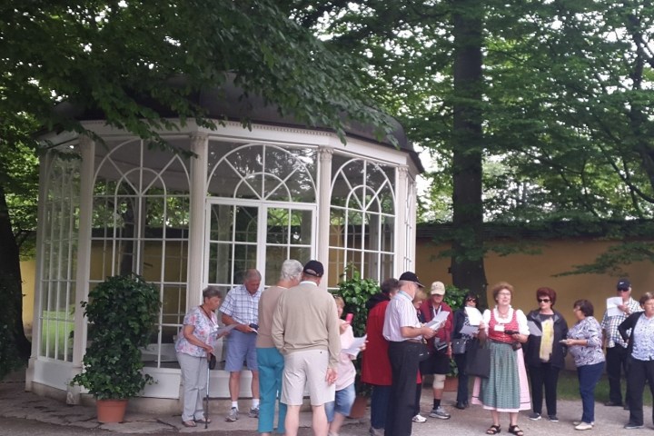 Group of people stand near a gazebo in a lush, green park setting.