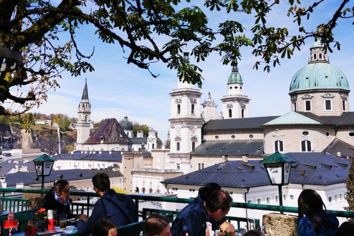People sitting on a terrace with a view of domed buildings and spires under a tree in a scenic cityscape.
