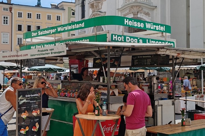 People standing at a sausage stand in a sunny outdoor market.