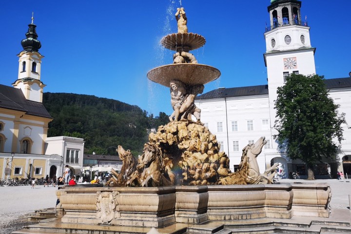Ornate fountain in a square, surrounded by historic buildings under a clear blue sky.