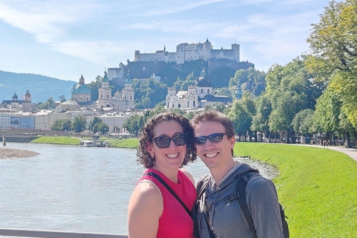 Two people smiling by a river, with a castle and cityscape in the background on a sunny day.