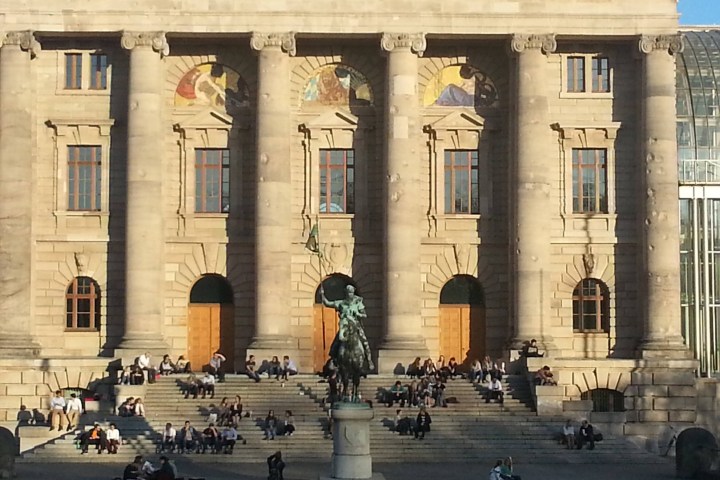 People sitting on steps in front of a large building with columns and a statue.