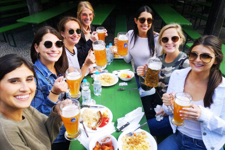 Group of women at outdoor table holding beer mugs, plates of food in front.