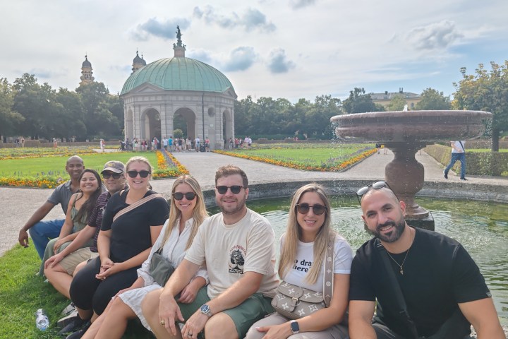 Group of people sitting by a fountain in a park with a domed pavilion in the background.
