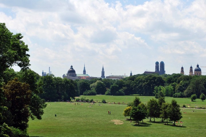Park scene with trees, open lawn, and a city skyline in the distance under a cloudy sky.