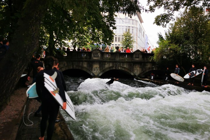 Surfer on river wave near bridge, surrounded by onlookers with surfboards.