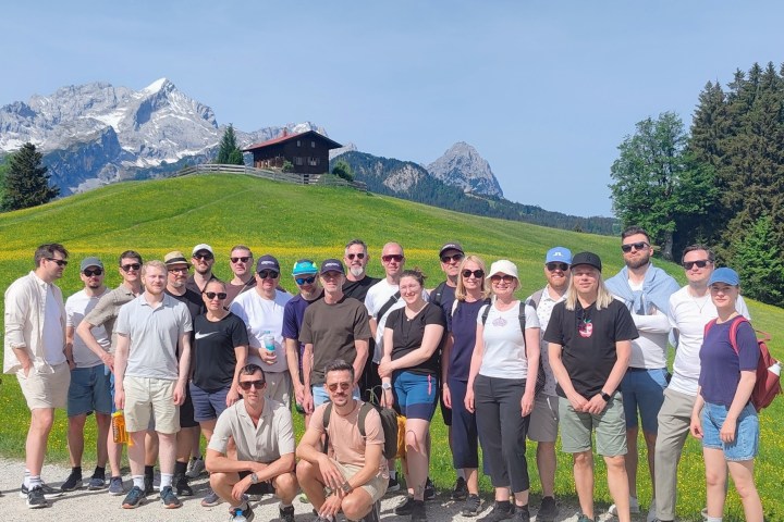 Group of people standing on a grassy hill with mountains in the background.