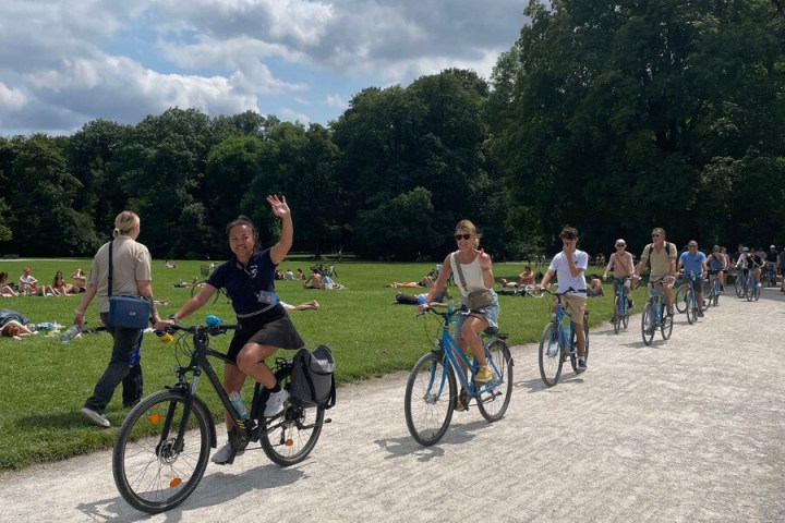 Group of people biking on a path beside sunbathers in a park.